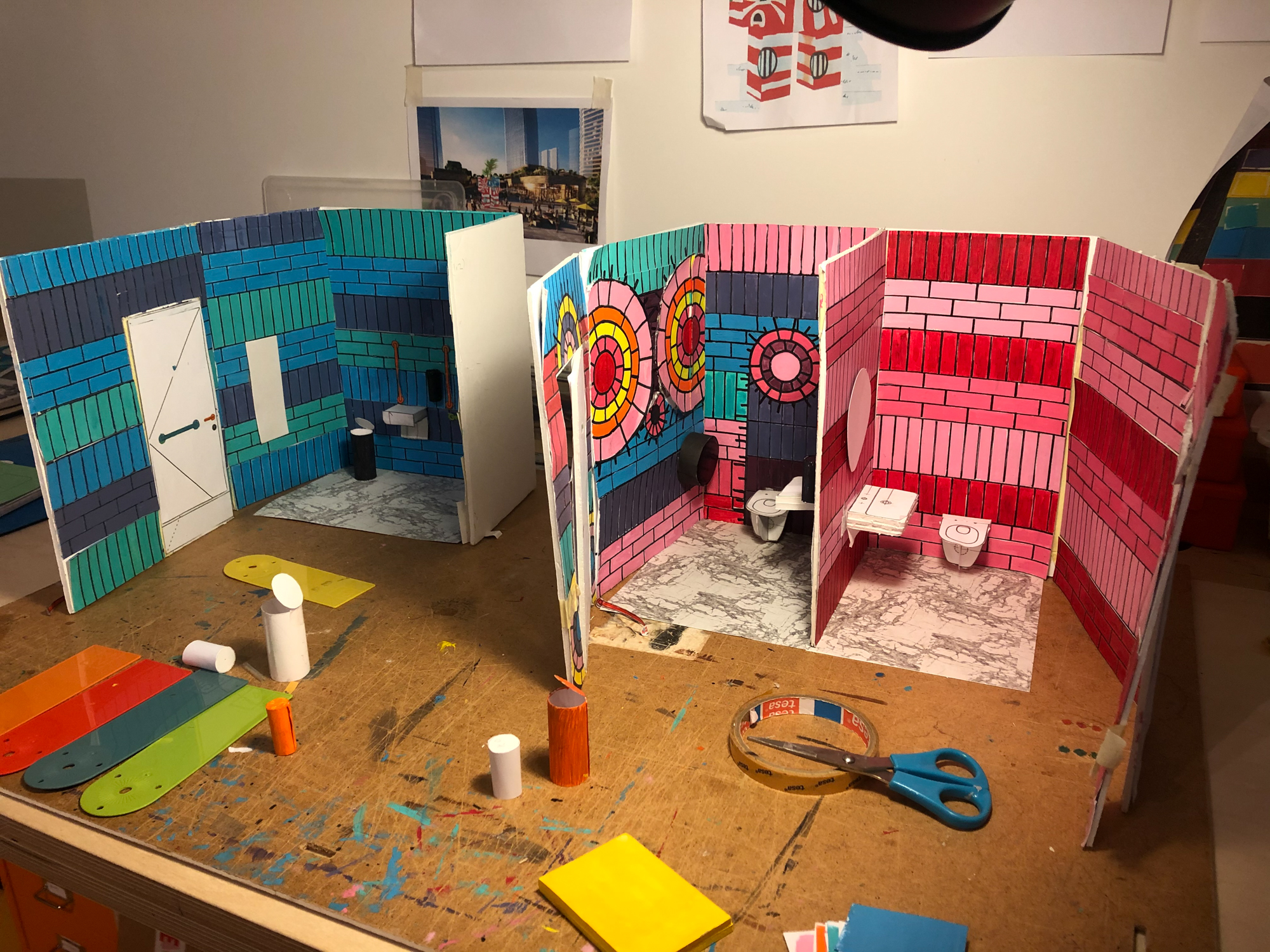 A photograph of 3 maquettes of the toilets on a brown desk. The maquettes show the blue, pink and green tiles, doors, sinks and toilets.
